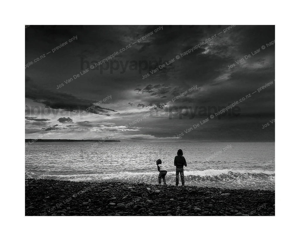 Two children standing at the edge of the sea on a rocky beach — Black & White photo by Jos Van De Laar