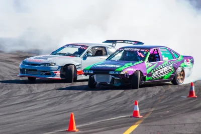 Two drift cars competing side by side on a sealed track with tyre smoke
